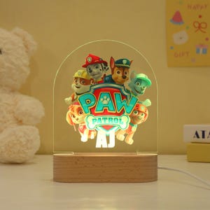 May include: A Paw Patrol night light featuring the show's logo and characters. The light is made of clear acrylic with a wooden base. The Paw Patrol logo is in teal and white, and the characters are in various colors. The base has the initials "AJ".