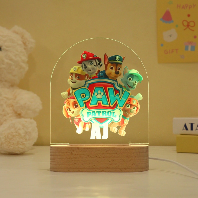 May include: A Paw Patrol night light featuring the show's logo and characters. The light is made of clear acrylic with a wooden base. The Paw Patrol logo is in teal and white, and the characters are in various colors. The base has the initials "AJ".