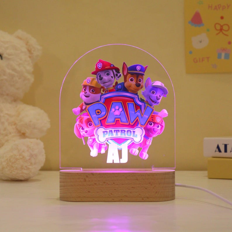 May include: A Paw Patrol night light featuring the show's characters, with the Paw Patrol logo in the center. The light is set on a wooden base and has a clear acrylic front. The light is powered by a white cord.