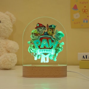 May include: A Paw Patrol night light featuring the show's characters, with the Paw Patrol logo in the center. The light has a wooden base and a clear acrylic panel with a green glow. The light is approximately 7 inches tall.