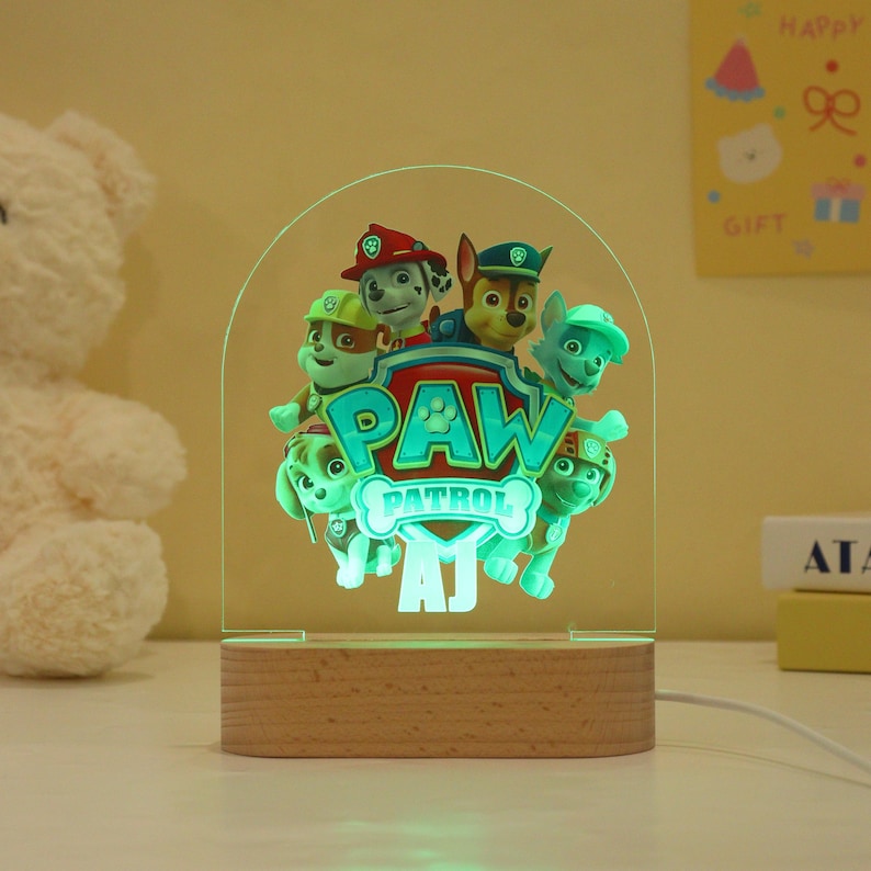 May include: A Paw Patrol night light featuring the show's characters, with the Paw Patrol logo in the center. The light has a wooden base and a clear acrylic panel with a green glow. The light is approximately 7 inches tall.