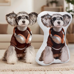 May include: A dog wearing a brown sweater and headphones, next to a dog-shaped pillow with the same image. The dog has gray and white fur and is wearing a brown sweater and headphones. The pillow is white with the dog's image printed on it.