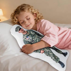 May include: A white pillow featuring a printed image of a person in military uniform. A young girl in a pink pajama top is holding the pillow. The pillow has an unusual shape.