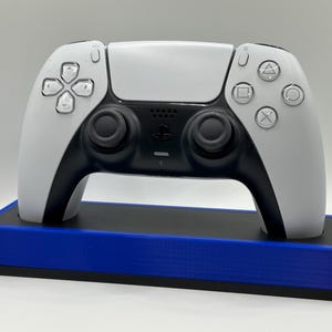 May include: A white and black PlayStation 5 controller resting on a blue and black charging stand. The controller features a symmetrical design with black joysticks and action buttons. The stand is rectangular and provides a stable base for the controller.