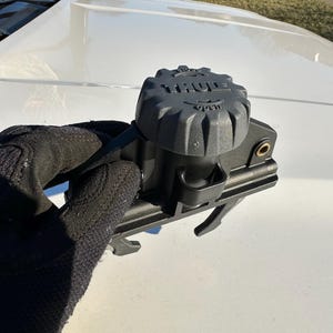 Puede incluir: Un componente negro de portaequipajes Thule con una parte superior texturizada y la palabra "THULE" en relieve. El componente tiene una perilla con la etiqueta "OPEN" y está sostenido por una mano enguantada. El portaequipajes tiene un soporte de montaje.