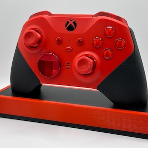 May include: A red Xbox controller with black grips rests on a matching red and black stand. The controller features a black Xbox logo and various buttons. The stand is rectangular and provides a stable base for the controller.