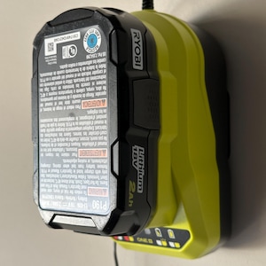 May include: A black and lime green Ryobi 18V Lithium 2Ah battery charger mounted on a wall. The charger features the Ryobi logo and warning labels. The charger is connected to a power outlet.