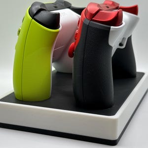 May include: A charging station for gaming controllers. The station is white with a black top and holds three controllers. The controllers are lime green, white, and black with red accents. The controllers are arranged in a row.