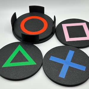 May include: Set of four black coasters with colorful game controller symbols: a red circle, pink square, green triangle, and blue X. The coasters are round and made of a textured material, with a holder for one coaster.