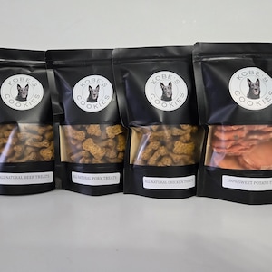 May include: Four black bags of dog treats with clear windows showing the treats inside. The bags are labelled "Kobe's Cookies" and have different flavours: beef, pork, chicken, and sweet potato.