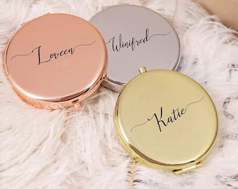 May include: Three personalized compact mirrors in rose gold, silver, and gold. Each round mirror has a name in elegant script: Loveen, Winifred, and Katie. The mirrors are displayed on a bed of white feathers.