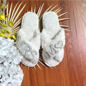 May include: A pair of white fuzzy slippers with the words "MRS" and "MR" embellished with rhinestones. The slippers are on a wooden surface.