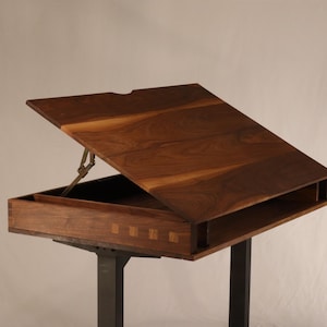 以下が含まれることがあります： 収納スペースを備えた、ヒンジ付きの天板を持つ木製の製図台。テーブルはウォールナット材で作られており、黒い金属製の脚が付いています。