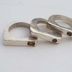 May include: Three silver rings with a square design are stacked on a white surface. Each ring has a small, coloured gemstone embedded in the band. One ring has a yellow stone, and the other two have red stones.