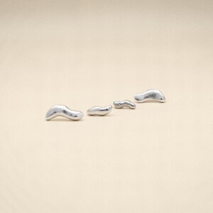 May include: Four small, silver-colored, abstract-shaped objects are arranged diagonally on a beige surface. The objects appear to be cast metal, with a smooth, reflective surface. The shapes are organic and irregular, resembling small, stylized blobs.