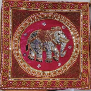 Könnte beinhalten: Ein rosa Samtkissen mit goldener Stickerei am Rand und einem weißen Elefantenmotiv in der Mitte. Der Elefant ist mit Pailletten und Perlen verziert.