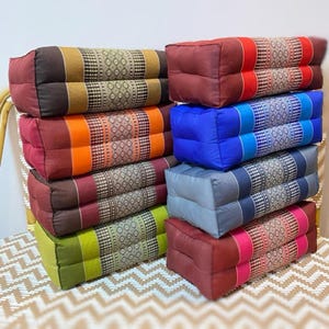 Pode incluir: Uma pilha de oito almofadas de meditação retangulares em várias cores e padrões, incluindo vermelho, azul, verde e marrom. As almofadas são feitas de tecido e têm um design tradicional tailandês.