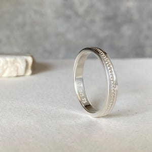 Könnte beinhalten: Ein silberner Ring mit einem Perlenmuster in der Mitte. Der Ring liegt auf einer weißen Oberfläche vor einem grauen Hintergrund. Im Inneren des Rings befindet sich eine kleine Inschrift.