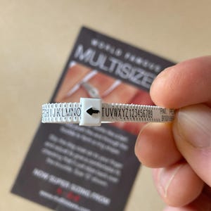 Può includere: Strumento di misurazione della taglia dell'anello in plastica bianca con lettere nere. Lo strumento è contrassegnato da lettere dalla F alla Z e da numeri da 1 a 9. Il testo "WORLD FAMOUS MULTISIZE" è visibile sullo sfondo.