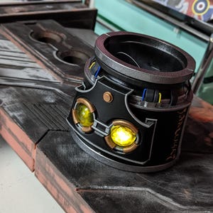 Puede incluir: Un objeto cilíndrico negro y plateado con un diseño decorativo. Tiene dos luces amarillas brillantes y un borde plateado. El objeto está sobre una superficie negra y marrón.