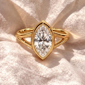 May include: A gold ring featuring a central marquise-cut clear gemstone set in a smooth bezel. The band has a split-shank design that tapers towards the centre stone, creating a sleek and polished look with a bright, reflective metallic finish.