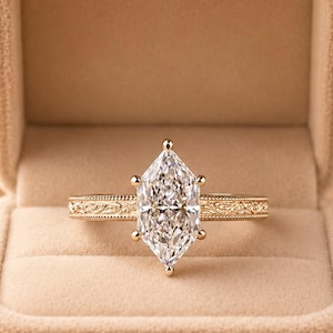 May include: A marquise-cut diamond ring set in a yellow gold band with intricate floral engraving along the shank. The ring is displayed inside a beige velvet jewelry box, featuring a single central stone held by six prongs in a classic setting.