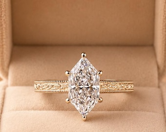 May include: A marquise-cut diamond ring set in a yellow gold band with intricate floral engraving along the shank. The ring is displayed inside a beige velvet jewelry box, featuring a single central stone held by six prongs in a classic setting.