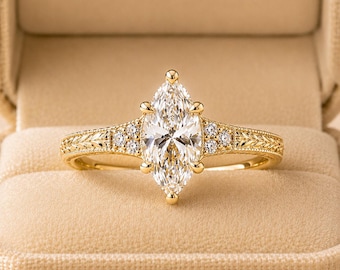 May include: A gold ring featuring a central marquise-cut clear gemstone held by six prongs. The band displays an intricate engraved leaf pattern on both sides, accented by three small round clear stones flanking the center setting.