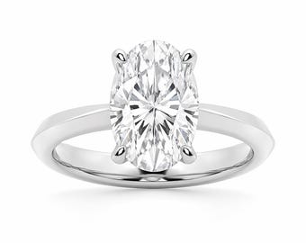 May include: A solitaire engagement ring featuring an oval-cut diamond set in a polished, white metal band. The diamond is held in place by four prongs, showcasing its brilliance. The ring is set against a clean white background.
