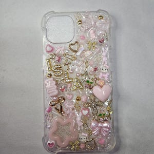May include: A clear phone case with a pink and gold glitter design. The case is decorated with various charms, including hearts, stars, bows, and the word "ISLA".