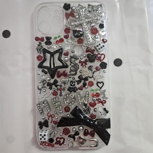 May include: A clear phone case decorated with black, red and silver embellishments. The case features a star, a cross, a bow, cherries, dice and the word "MEOW" in rhinestones.