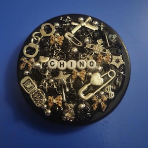 May include: A black, round decorative piece adorned with various charms, including handcuffs, safety pins, stars, and bows. The word "CHINO" is spelled out in white letter beads. The design incorporates silver, black, and leopard print accents.