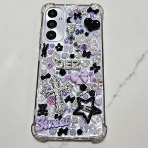 Peut inclure: Une coque de téléphone transparente avec un design violet, noir et argent. La coque est recouverte de strass, de breloques et d'autres embellissements. Le texte "Sweet" et "DEZ" est visible sur la coque.