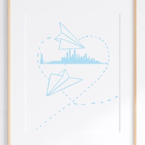 May include: Framed print with a light blue illustration of two paper aeroplanes forming a heart shape around a city skyline. The artwork includes the Statue of Liberty and a cityscape.