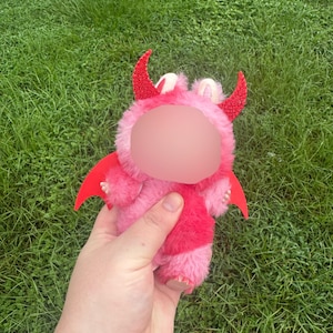 May include: A pink plush toy in the shape of a devil, with red horns, wings, and a red chest. The toy is held in a hand against a background of green grass.