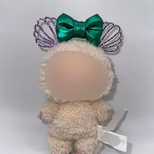 May include: A plush toy in the shape of a small creature, with a fluffy, cream-colored body. It has a green satin bow and purple beaded accents on its head. The toy has pink feet and a tag attached.