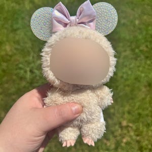 May include: A plush toy character with a light beige body and pink feet. The toy has a white fluffy head with a light purple bow and sparkly ears. The toy is held against a green grassy background.