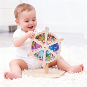 May include: A wooden baby toy with a wheel design. The wheel has six sections with colorful illustrations and small beads. The toy is on a wooden base and is being held by a baby wearing a white onesie.