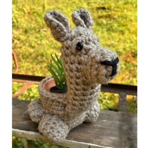 Puede incluir: Un macetero de llama tejido a crochet en color marrón claro. El macetero tiene una maceta pequeña en el interior para una planta. El macetero de llama está sentado sobre una superficie de madera. El texto "Llama Planter" está en la parte inferior de la imagen.