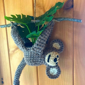 May include: A crocheted monkey hanging from a branch with a green plant inside. The monkey is brown with a beige face and ears.