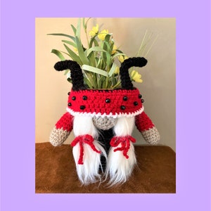 May include: A crocheted gnome wearing a red and black ladybug hat with black bead accents. The gnome has white hair and red bows tied in the hair. The gnome is holding a potted plant with green leaves and yellow flowers.