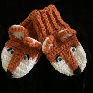 May include: Crocheted fox-shaped baby booties in orange, white, and black. The booties feature fox faces with googly eyes and black noses. The ribbed cuff and fox head are orange, with white accents on the face.