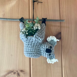 Puede incluir: Un koala de crochet sosteniendo una maceta pequeña con flores blancas colgando de una rama de madera. El koala es gris con ojos negros y pelaje blanco en las orejas.