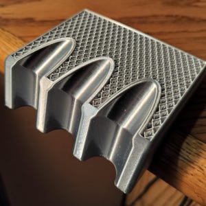 Puede incluir: Un cenicero plateado con un patrón de diamantes y tres apoyos en forma de bala. El cenicero es de metal y tiene un diseño moderno y geométrico. El cenicero se muestra sobre una superficie de madera.