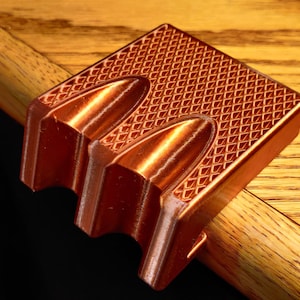 Puede incluir: Un objeto de color cobre impreso en 3D con una superficie texturizada y tres ranuras redondeadas. El objeto está diseñado para ser utilizado como un soporte de teléfono o un organizador de escritorio.