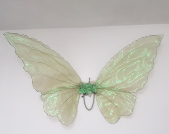 May include: Iridescent green butterfly wings with a central green floral accent. The wings are made of a sheer, shimmering fabric and are attached to a metal frame. The wings are designed to be worn or used as a decorative item.