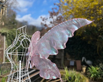 May include: A pair of iridescent pink fairy wings with a delicate, layered design. The wings are displayed on a white mannequin in a garden setting.