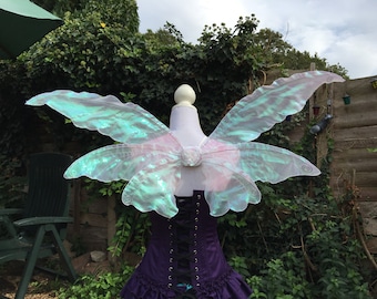 May include: A pair of iridescent fairy wings with a light blue and green color scheme. The wings are made of a sheer, translucent material and are attached to a white base.