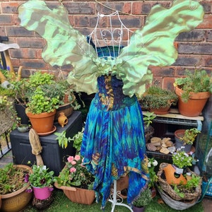 May include: A whimsical fairy costume displayed on a white wire dress form. The costume features iridescent green wings and a blue and gold patterned dress. The background includes potted plants and a brick wall, creating an enchanting outdoor scene.