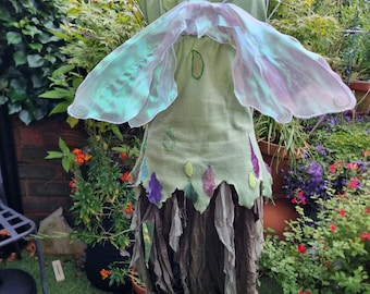 May include: A light green fairy costume with iridescent wings, displayed on a white mannequin. The dress features colorful leaf accents and a layered, brown skirt. The background includes lush greenery and potted plants.
